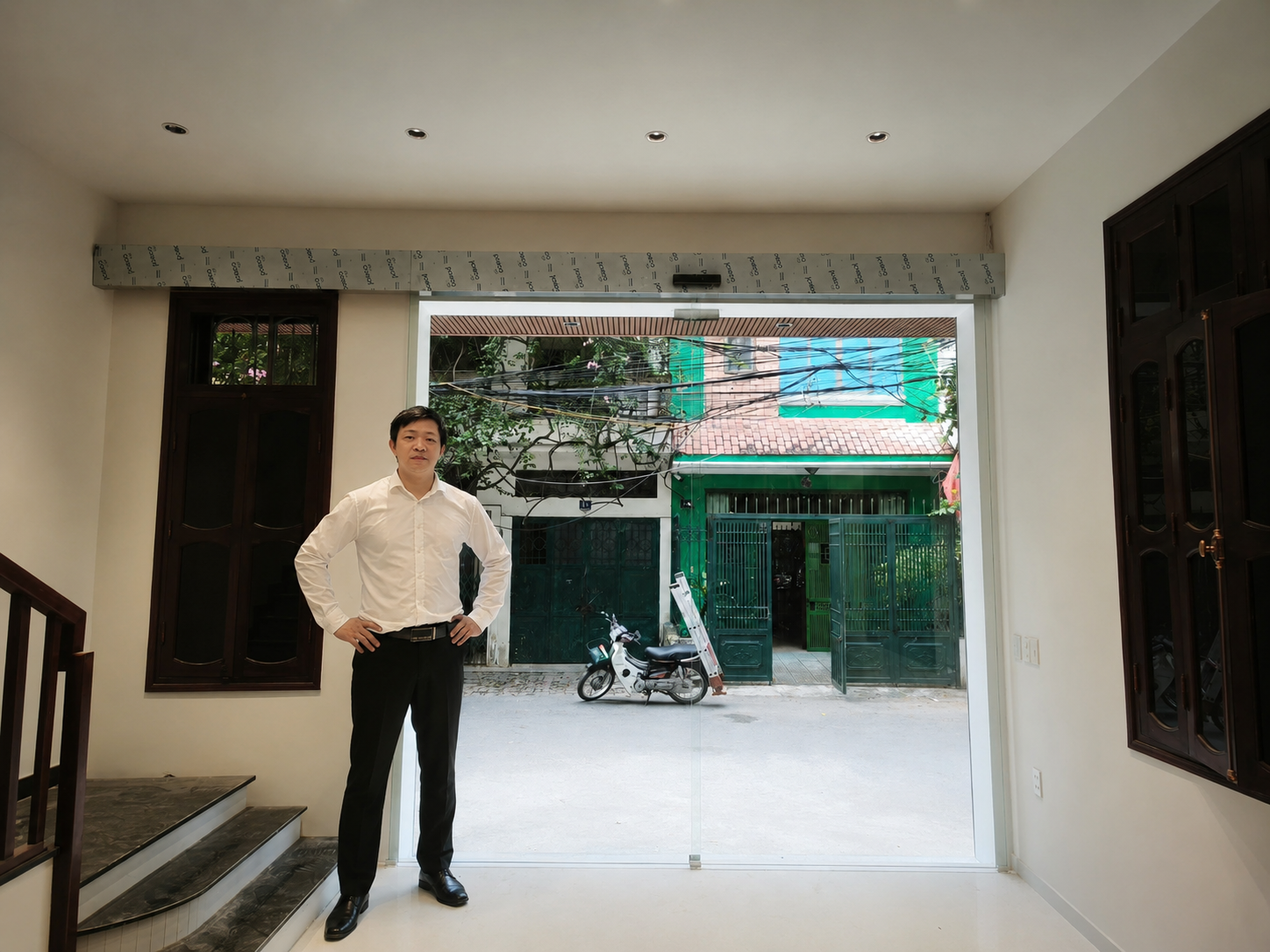 Người đàn ông mặc sơ mi trắng quần âu đen đứng trong nhà mới với cửa kính lớn nhìn ra phố
